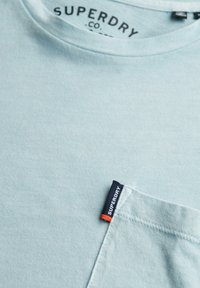 Camiseta azul claro con cuello redondo y bolsillo. Presenta una etiqueta de marca negra y naranja en el bolsillo, fabricada con suave tela de algodón.