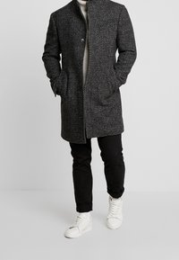 Grå mønstret frakke med opretstående krave, to frontlommer og knapper, parret med sorte bukser og hvide sneakers.