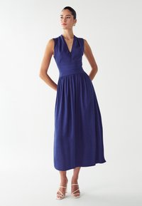 Mornarskoplavi haljina bez rukava s V-izrezom, tijesnim gornjim dijelom i lepršavom suknjom do koljena. Model nosi bijele sandale s trakama.