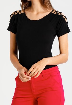 Woman wearing a black short-sleeve top with crisscross shoulder cutouts and bright red pants, hands clasped in front.
