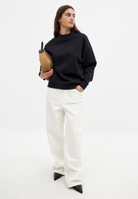 Czarny oversize'owy sweatshirt z wysokim kołnierzem. W połączeniu z białymi szerokimi spodniami i czarnymi butami na szpilce. Trzyma brązową torbę.