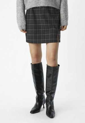 Person wearing a gray plaid mini skirt, gray sweater, and knee-high black leather boots with heels, standing against a white background.