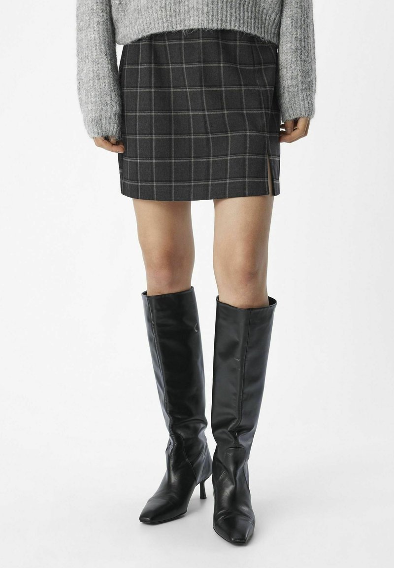 Personne portant une mini-jupe à carreaux grise, un pull gris et des bottes en cuir noires montantes à talons, debout sur un fond blanc.