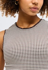 Άτομο με μπούκλες φοράει χρυσό κρίκο σκουλαρίκι και ασπρόμαυρο καρό τοπ χωρίς μανίκια με βολάν στις άκρες.