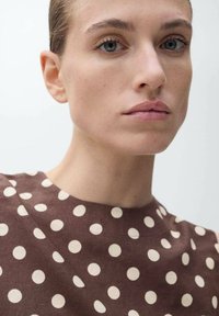 Hnědý lněný top s kulatým výstřihem, který má velké kr�émové puntíky a texturovaný povrch. Pleť vypadá přirozeně a nezkrášleně.