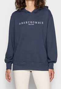 Granatowa bluza z kapturem, wykonana z miękkiego materiału, z kieszenią z przodu, kapturem ze ściągaczem i napisem "ABERCROMBIE New York" w kolorze białym na klatce piersiowej.