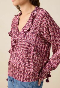 Frau, die eine lila blumige Bluse mit Rüschen und Quasten an den Ärmeln trägt, kombiniert mit blauen Jeans, seitlich gezeigt.