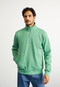 Pullover de fleece verde com fecho, padrão texturizado, gola alta e bolsos frontais. Projetado para uso casual com um corte descontraído.