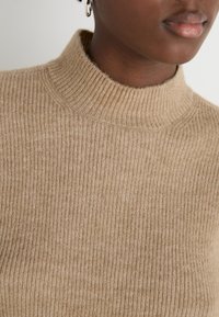 Knit turtleneck sweater in light brown, featuring a ribbed texture and fitted design. Close-up view highlights the neckline and fabric details.
