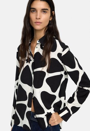 Kobieta o długich ciemnych włosach, ubrana w czarno-białą koszulę w guziki z wzorem oraz niebieskie jeansy, z jedną ręką na biodrze.