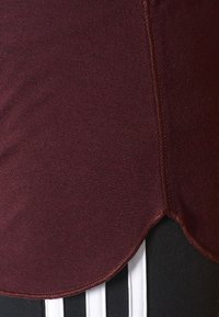 Bordeauxfärgad idrottstopp med slät tyg, rundad nederkant och kontrasterande svarta ränder nedtill. Synlig sömdetalj förbättrar designen.