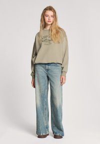 Oliwkowy sweatshirt z graficznym nadrukiem, w połączeniu z jasnoniebieskimi szerokimi spodniami dżinsowymi. Miękka tkanina, luźny krój i wyblakła faktura dżinsu.