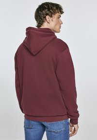 Burgunderfarbener Pullover-Hoodie mit Kängurutasche, verstellbarem Kordelzug sowie gerippten Bündchen, kombiniert mit blauen Jeans.