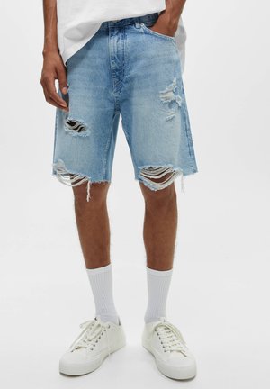 Lysblå denimshorts med frynsede kanter og flere store rifter. Har en klassisk pasform med høj talje og bæltestropper. Bæret med hvide sneakers.