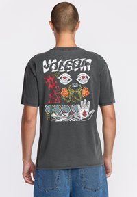 T-shirt gris avec un grand motif coloré au dos, comprenant des yeux, des flammes, des fleurs et divers motifs, fabriqué en tissu coton doux.