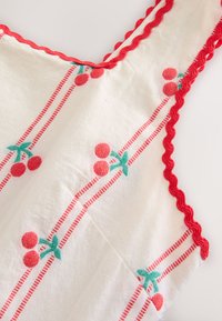 Bílá látka s vertikálními červenými pruhovanými vzory třešní a vroubkovaným červeným lemováním podél okraje bezrukávového oděvu.