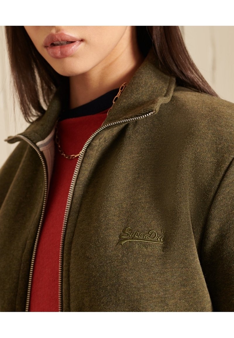 Veste zippée vert olive avec une finition texturée, dotée d'un logo en relief et d'une couche inférieure rouge contrastante. Détail de fermeture éclair lisse.