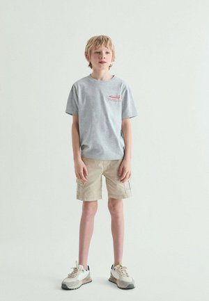 Niño rubio de pie, con una camiseta gris con estampado rojo, pantalones cortos cargo beige y zapatillas blancas, sobre un fondo liso y claro.