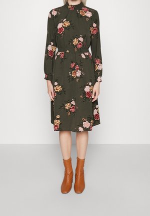 Vrouw die een lange mouwen olijfgroene jurk draagt met roze en gele bloemenpatronen, gecombineerd met beige enkellaarzen, staand op een lichte vloer.
