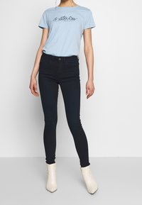 Camiseta de manga corta azul claro con texto negro, combinada con jeans ajustados azul oscuro y botines crema. Diseño sencillo, tela suave.