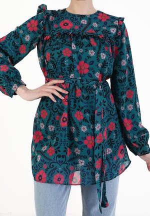 Femme portant un chemisier à manches longues couleur sarcelle avec un motif floral rouge et rose, volants sur la poitrine et ceinture nouée à la taille, associé à un jean bleu clair.