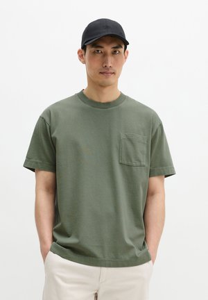 Mann mit schwarzer Kappe, olivgrünem T-Shirt mit Tasche und hellbeigen Hosen, steht mit Händen in den Taschen vor einfachem weißem Hintergrund.