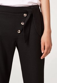 Pantaloni neri a vita alta con cintura annodata, dotati di bottoni argentati sui lati e una superficie di tessuto liscia.