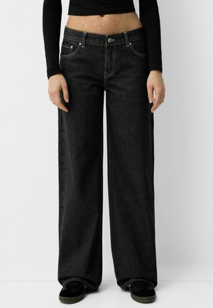 Person wearing black wide-leg jeans with visible stitching, paired with black long-sleeve crop top and black shoes against a plain background.