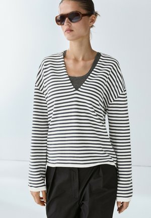 Frau mit brauner Sonnenbrille, weiß-schwarz gestreiftem Langarmshirt über einem dunklen gerippten Hemd und schwarzen Hosen, stehend vor weißem Hintergrund.