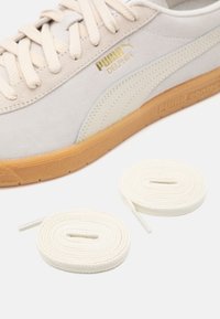 Beige Puma Dolphin-sneaker med gummisula, guldlogotyp och två rullade vita skosnören bredvid den på en vit yta.