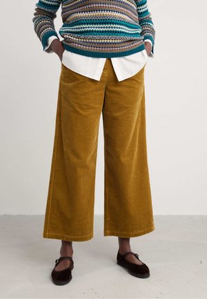 Mustard corduroy wide-leg pants with vertical ribbing, paired with a white collared shirt under a multi-colored patterned sweater.