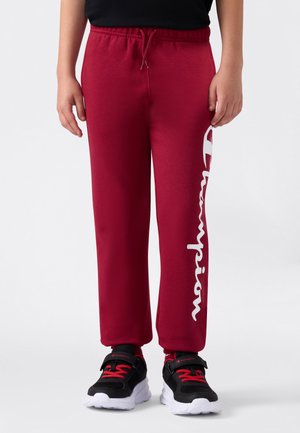 Kind in rode sweatpants met het witte 'Champion' logo op het linkerbeen en zwarte sneakers met rode veters op een witte achtergrond.