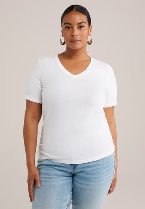 Femme aux cheveux noirs portant un T-shirt blanc à col V et des jeans bleu clair, regardant de face contre un fond uni.