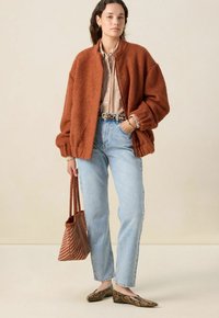 Veste bomber en polaire rouille avec des manches surdimensionnées, chemise à rayures à boutons, jeans bleu clair, ballerines en cuir serpent et un sac fourre-tout tressé marron.