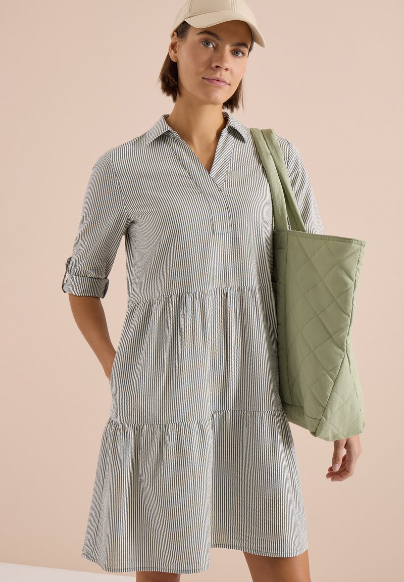 Femme portant une robe beige à rayures et une casquette crème, portant un sac cabas matelassé vert clair à l'épaule sur fond beige.