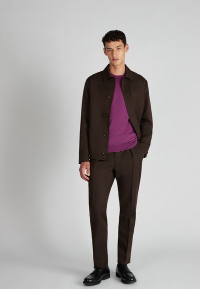 Giacca di tessuto marrone con bottoni neri sopra una camicia viola, abbinata a pantaloni marroni coordinati e scarpe nere. Design minimalista e vestibilità.