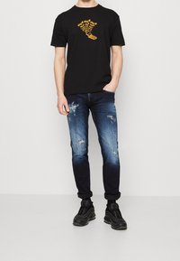 Svart kortärmad t-shirt med gul grafisk tryck, kombinerad med slitna blå denimjeans. Skor: svarta sneakers med texturerad detalj.