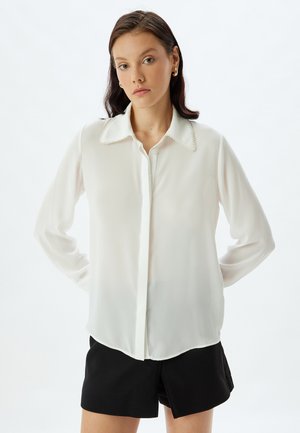 Blusa blanca y semitransparente con un cuello clásico que presenta un ribete decorativo, mangas largas y un cierre frontal con botones, combinada con pantalones cortos negros.