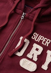 Felpa con zip color bordeaux con ricamo bianco "SUPERDRY 54". Presenta una zip in metallo e un cappuccio regolabile con cordino. Realizzata in cotone misto.