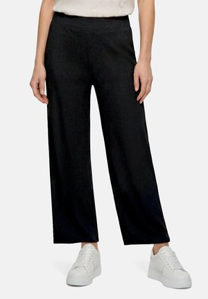 Personne portant un pantalon noir ample et des baskets blanches, debout avec les mains détendues le long du corps sur un fond uni.