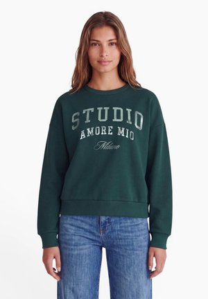 Kvinde med langt brunt hår iført en mørkegrøn sweatshirt med "STUDIO AMORE MIO Milano" tekst og blå jeans på hvid baggrund.