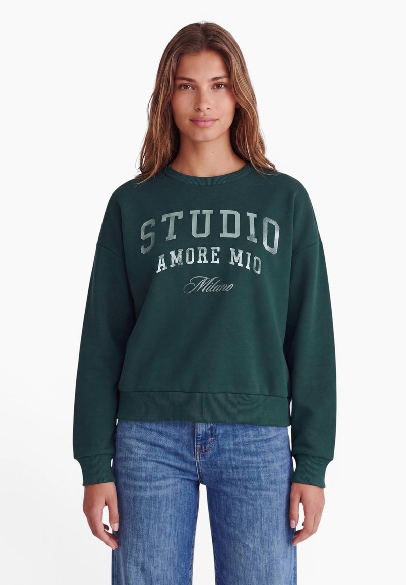 Femme aux longs cheveux bruns portant un sweat-shirt vert foncé avec le texte "STUDIO AMORE MIO Milano" et un jean bleu sur fond blanc.