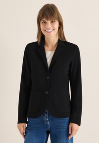 Schwarzer Blazer aus Stretchstoff, ausgestattet mit einem Reverskragen, zwei vorderen Taschen und einer Zwei-Knopf-Verschluss. Getragen über einem hellen Oberteil.