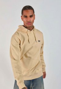 Un homme portant un hoodie beige avec une poche avant et un jean bleu se tient devant un fond clair et uni.