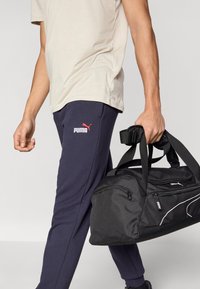 Μαύρη τσάντα τύπου σακ βουαγιάζ με φερμουάρ και λευκές λεπτομέρειες, που κρατιέται μαζί με ναυτικό παντελόνι φόρμας με κόκκινο λογότυπο Puma και μπεζ μπλουζάκι.