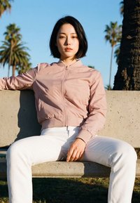 Young woman with short black hair wearing a pink jacket and white pants sitting on a concrete bench outdoors near palm trees.