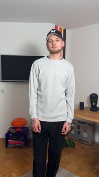 Felpa grigia a maniche lunghe con trama a coste, dettaglio logo sul petto, abbinata a pantaloni neri, in piedi in una stanza con mobili.