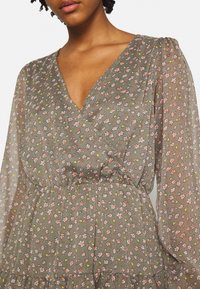 Robe à fleurs en tissu léger et transparent. Elle présente un décolleté en V, des manches longues, et une base verte avec de petites fleurs roses et jaunes.