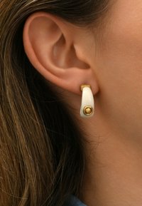 Boucles d'oreilles en céramique blanche avec un design courbé, présentant un accent doré à la base et une embellissement circulaire doré à l'avant.