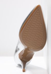 Chaussure à talon haut en métal argenté avec un bout pointu, présentant une semelle texturée couleur fauve avec un motif de logo MK et un talon fin.